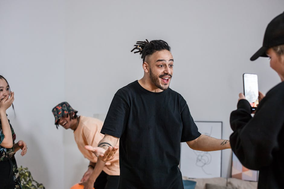 Smiling man in a black t-shirt performing while a friend records him on a smartphone.